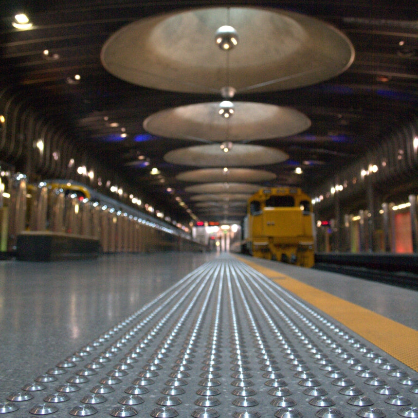 Low angle view of TacPro stainless steel tactile warning indicators on concrete platform edge. Moody ambient ceiling lighting. Britomart Train Station