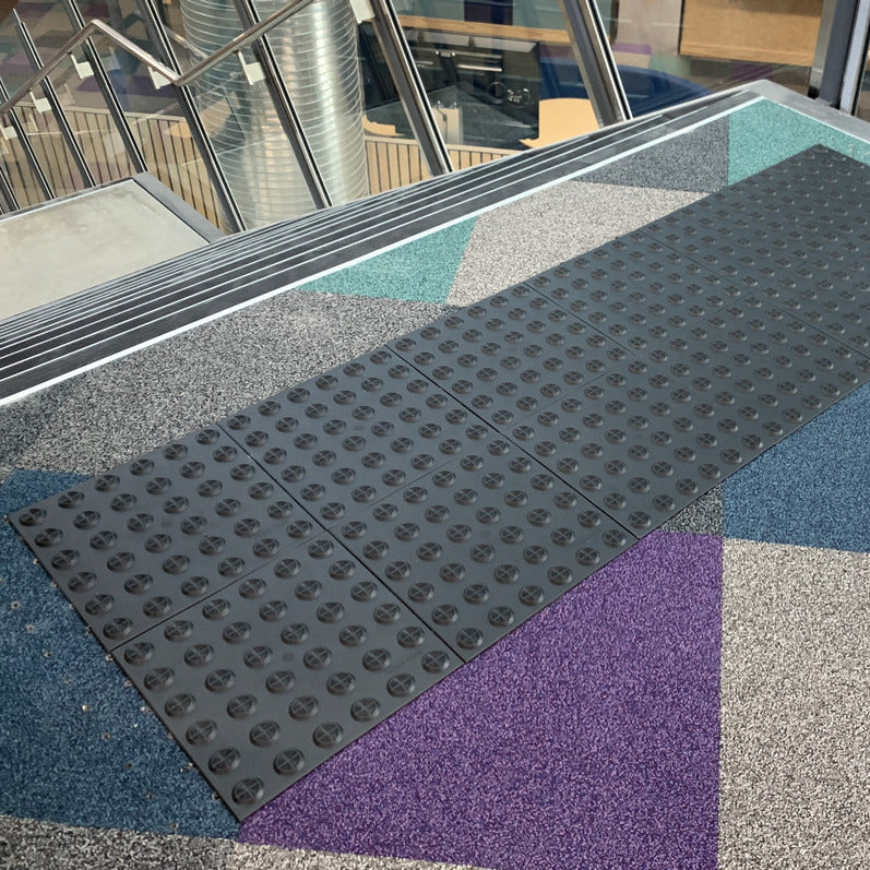 TacPro black peel-and-stick warning tactile tiles installed on multi-coloured carpet at the top of stairs at a school.